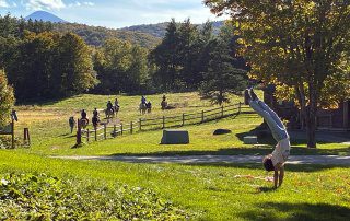 A student does a handstand in the grass. Horses can be seen riding away in the distance.
